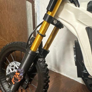 Puede incluir: Primer plano de una mini moto el&eacute;ctrica. La rueda delantera y la suspensi&oacute;n son visibles, con amortiguadores dorados y un neum&aacute;tico negro. La moto tiene un cuadro blanco y una horquilla negra. El logotipo de Fox es visible en la horquilla delantera.