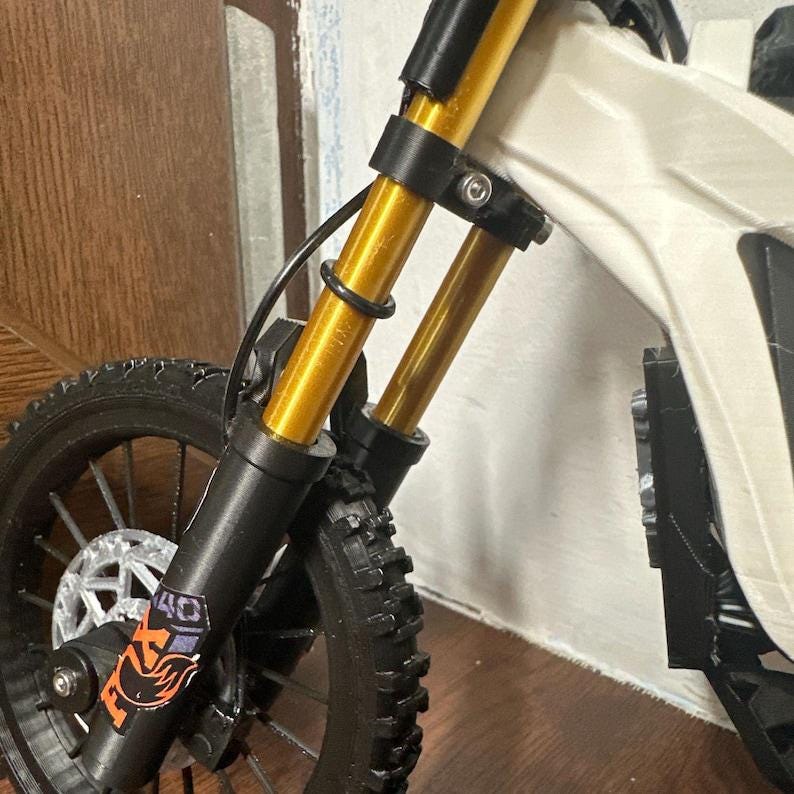 Puede incluir: Primer plano de una mini moto el&eacute;ctrica. La rueda delantera y la suspensi&oacute;n son visibles, con amortiguadores dorados y un neum&aacute;tico negro. La moto tiene un cuadro blanco y una horquilla negra. El logotipo de Fox es visible en la horquilla delantera.