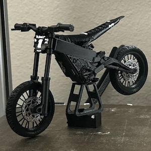May include: A black miniature motorcycle with a unique design, featuring a prominent front wheel and a raised rear wheel. The model includes detailed components like the handlebars, seat, and engine, and is supported by a black stand. The front of the motorcycle has the number "1".