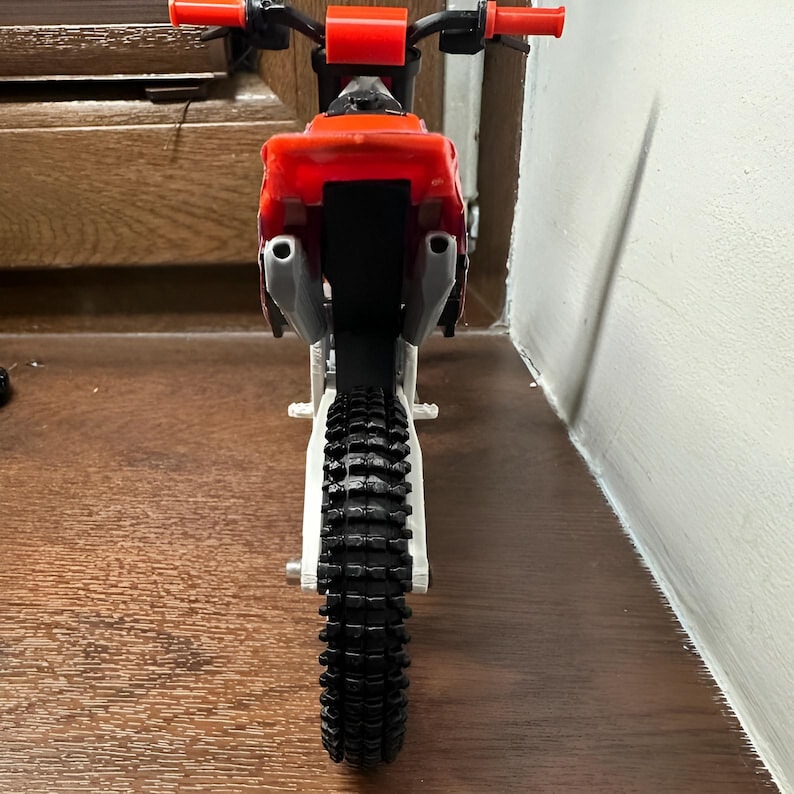 May include: A miniature toy dirt bike, viewed from the rear. The bike features a red body, black tires, and a white frame. The handlebars and exhaust pipes are visible. The tires have a deep tread pattern.