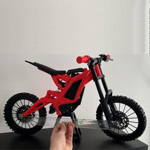 K&ouml;nnte beinhalten: Ein rotes und schwarzes Miniaturmotorrad mit schwarzen Reifen und einem schwarzen Sitz. Der Lenker und die Griffe sind rot. Das Motorrad steht auf einer schwarzen Oberfl&auml;che. Ein Lineal wird in der N&auml;he des Vorderrads gehalten. Das Motorrad ist etwa 30 cm lang.