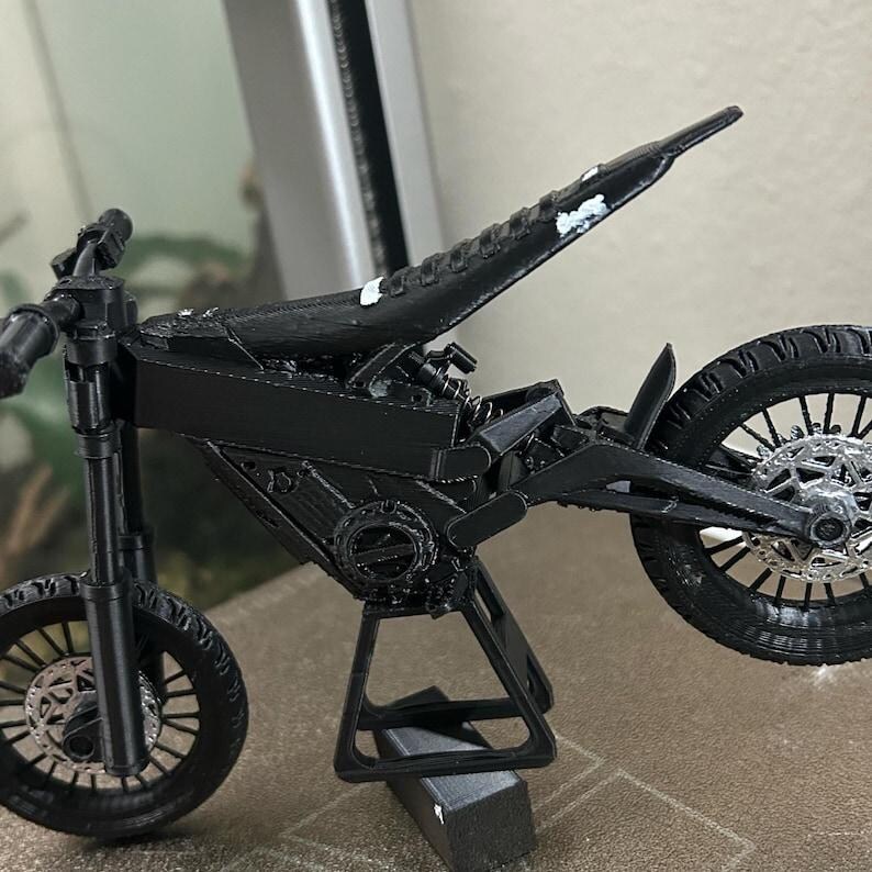 Puede incluir: Modelo de motocicleta en miniatura negra con neum&aacute;ticos, ruedas y marco detallados. El modelo est&aacute; soportado por un soporte negro, mostrando el intrincado dise&ntilde;o del veh&iacute;culo. El asiento tiene pintura blanca.