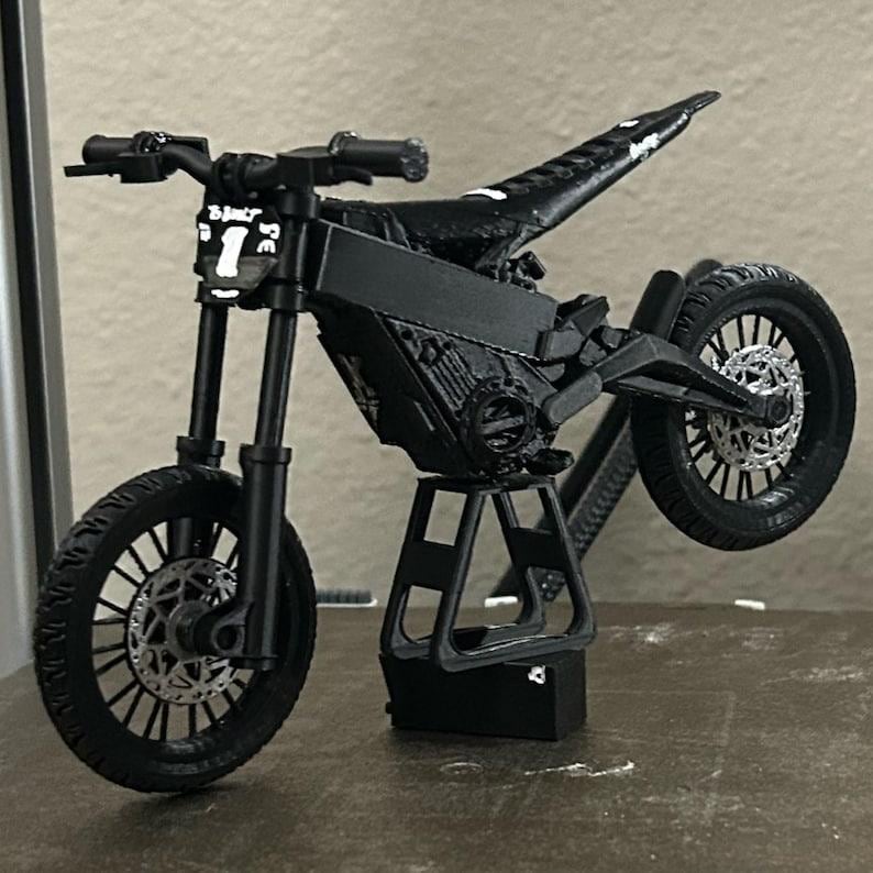 Może przedstawiać: Czarny model motocykla o unikalnym designie, z wyraźnym przednim kołem i podniesionym tylnym kołem. Model ma skomplikowane detale, w tym kierownicę, siedzenie i elementy silnika. Z przodu motocykla widnieje numer "1".