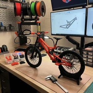 Pu&ograve; includere: Un modello di mountain bike rosso e nero &egrave; esposto su un banco da lavoro, con un calibro digitale in primo piano. La bici si trova in un'officina con attrezzature per la stampa 3D e bobine di filamento. Un monitor di computer mostra un design del telaio della bici.