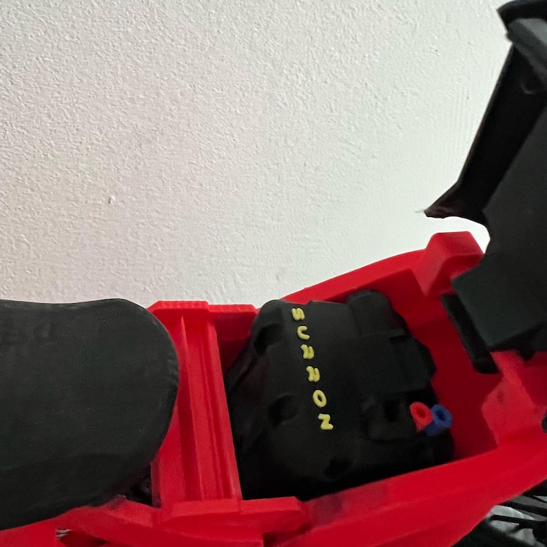 May include: A red electric motorcycle frame with a black battery pack. The battery pack has the word "SURRON" in yellow lettering. The motorcycle seat is black. The motorcycle is against a white wall.