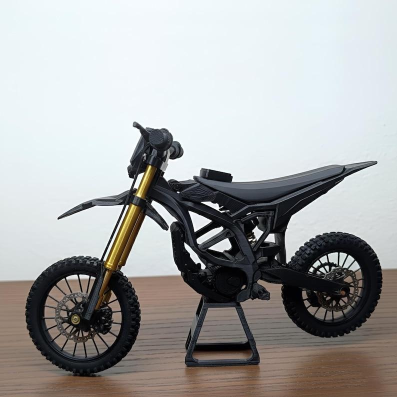 May include: A black and gold miniature dirt bike model. The model features a black frame, tires, and seat, with gold-colored front forks. The bike is displayed on a small black stand, showcasing its design and details.
