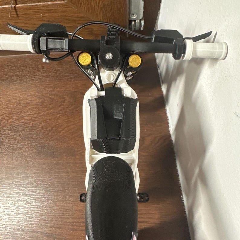 May include: A close-up view of a small, white and black scooter. The handlebars are black with white grips. The scooter has yellow accents and a black front wheel. The scooter is on a wooden surface.