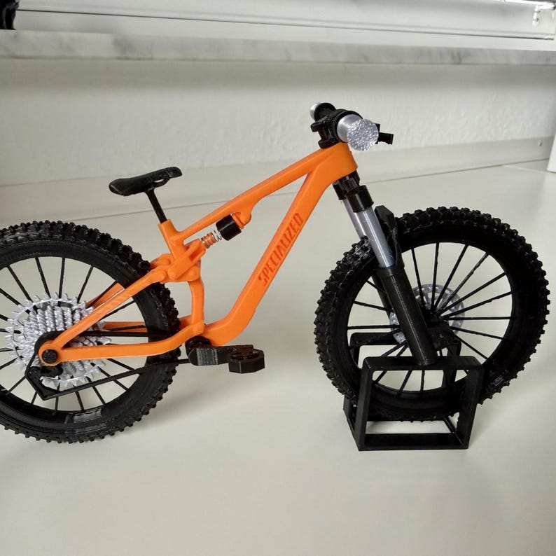 May include: An orange miniature mountain bike with black tires and a black stand. The bike has the word "SPECIALIZED" printed on the frame. The bike has a black seat and pedals.