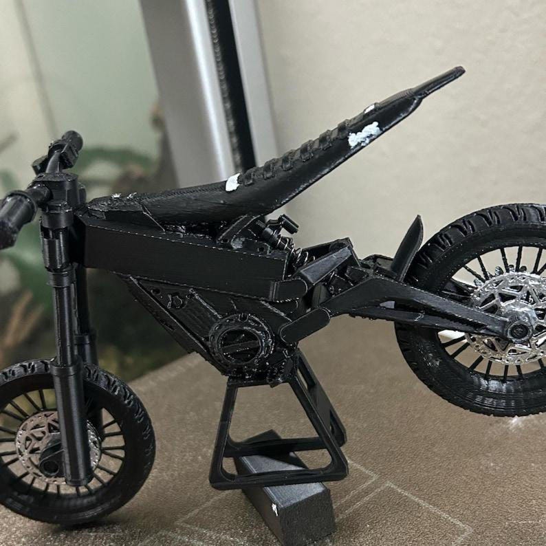 May include: A black motorcycle model with a unique design, featuring detailed wheels, a seat, and a stand. The model is made of a matte black material, showcasing the intricate design of a futuristic motorcycle.