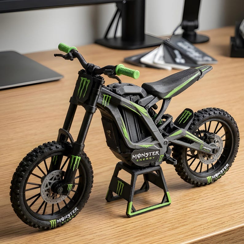 May include: A black and green toy motorcycle with the Monster Energy logo. The model has green accents on the handlebars, frame, and wheels. The tires are black with a tread pattern, and the motorcycle is on a black stand.