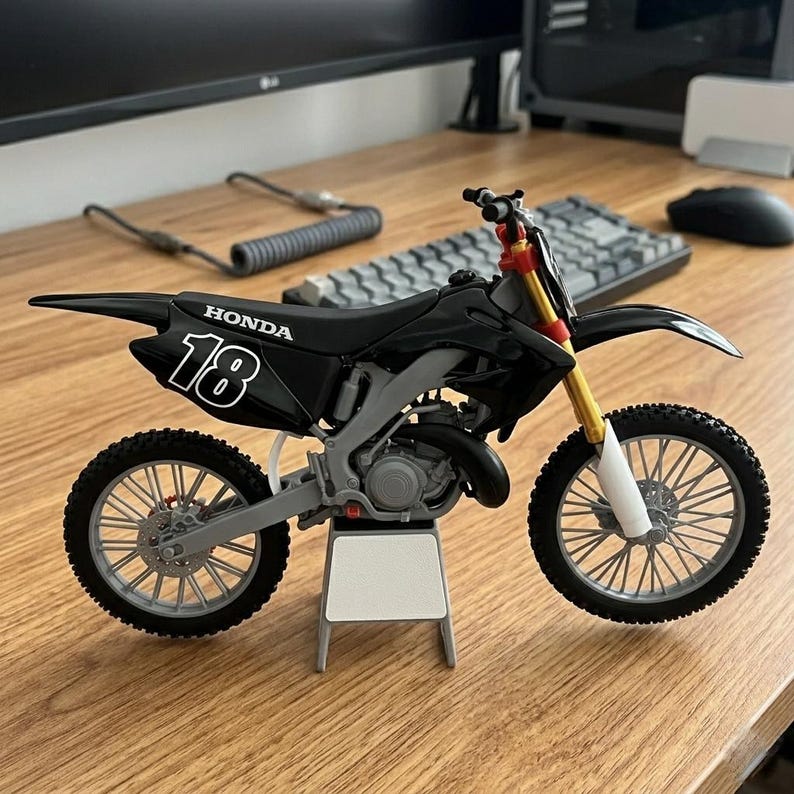 May include: A black and gray Honda model motorcycle with the number 18 on the side. The motorcycle has gold and red accents on the front forks and is resting on a small white stand. The tires are black with gray spokes.
