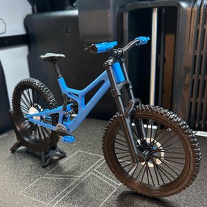 Pu&ograve; includere: Una mountain bike blu in miniatura con pneumatici neri e un telaio nero. La bici ha un design dettagliato, tra cui sospensioni, pedali e manubrio. La bici &egrave; esposta su una superficie scura.