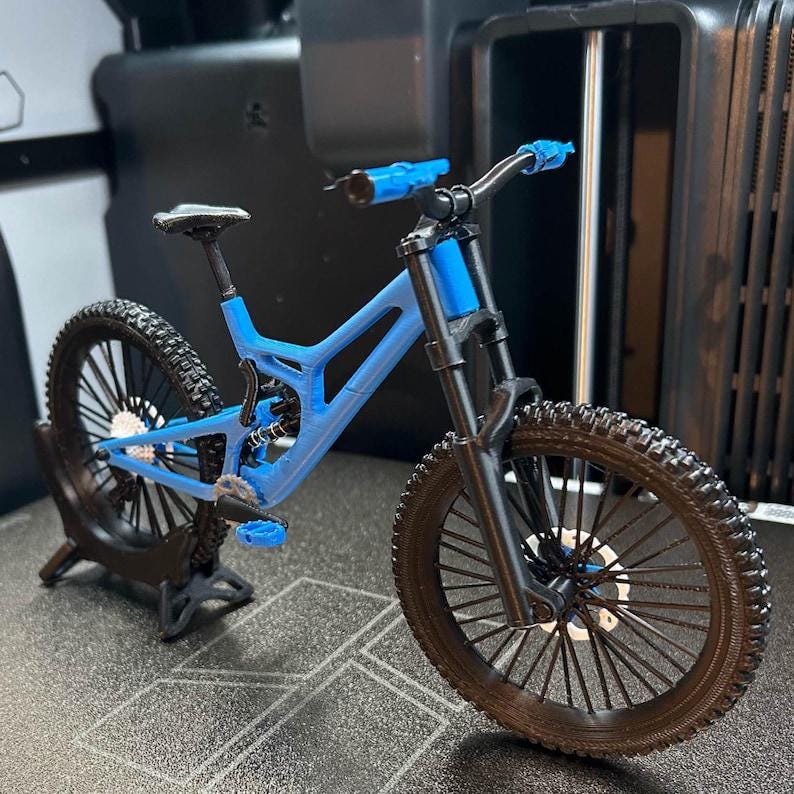 Pu&ograve; includere: Una mountain bike blu in miniatura con pneumatici neri e un telaio nero. La bici ha un design dettagliato, tra cui sospensioni, pedali e manubrio. La bici &egrave; esposta su una superficie scura.
