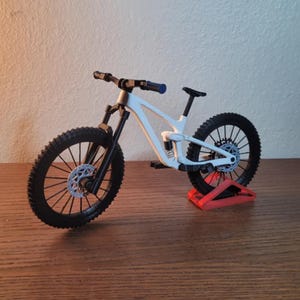 May include: A white and black mountain bike model with black tires and a blue handlebar grip. The bike is displayed on a red stand, showcasing its design and components.