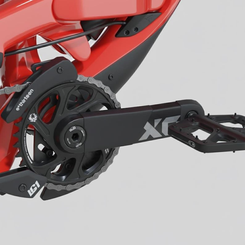 May include: Close-up of a red and black mountain bike frame with a black chainring, chain, and pedal. The chainring has the text "e*thirteen" and "LG1" printed on it. The pedal arm has the text "X" printed on it.