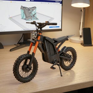 K&ouml;nnte beinhalten: Ein kleines schwarzes und orangefarbenes Elektromotorradmodell mit Stollenreifen. Das Modell hat einen Scheinwerfer, einen Sitz und einen St&auml;nder. Das Motorrad wird auf einer Holzoberfl&auml;che ausgestellt.