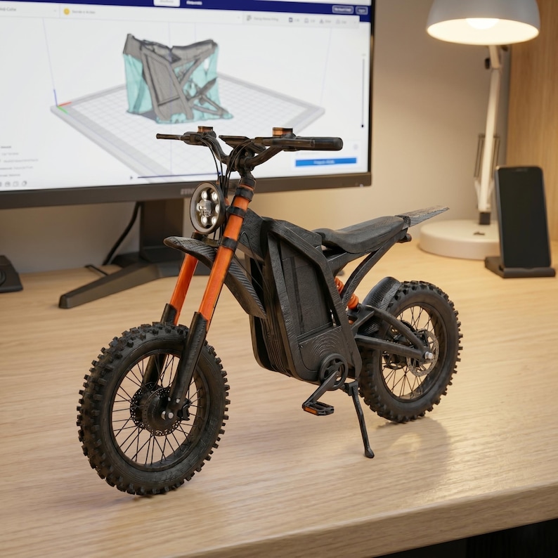 K&ouml;nnte beinhalten: Ein kleines schwarzes und orangefarbenes Elektromotorradmodell mit Stollenreifen. Das Modell hat einen Scheinwerfer, einen Sitz und einen St&auml;nder. Das Motorrad wird auf einer Holzoberfl&auml;che ausgestellt.