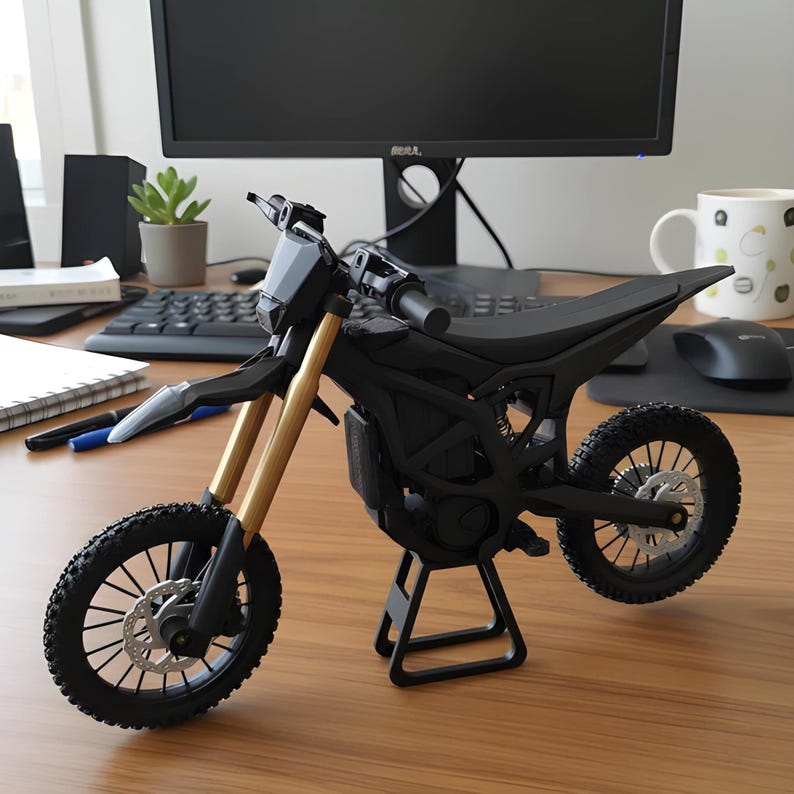 May include: A black and gold miniature motorcycle model with knobby tires and a black seat. The model has gold-colored front forks and is displayed on a black stand. The motorcycle is on a wooden desk with a computer monitor in the background.