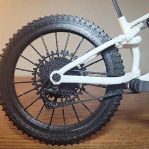 May include: Close-up of a black and white mountain bike rear wheel. The black tire has a textured tread. The wheel's spokes and gears are black, and the frame is white. The bike appears to be a miniature model.