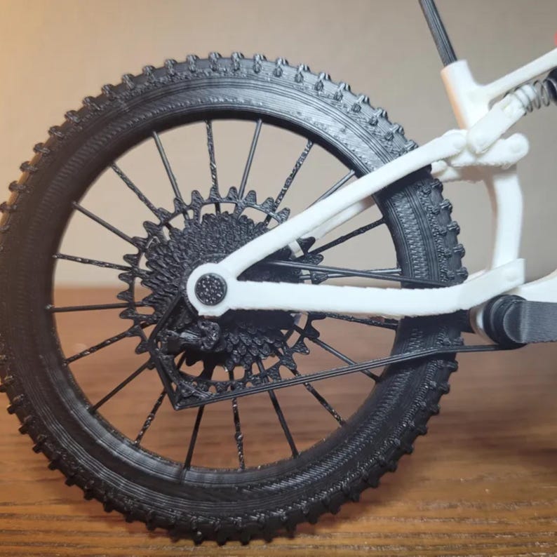 May include: Close-up of a black and white mountain bike rear wheel. The black tire has a textured tread. The wheel's spokes and gears are black, and the frame is white. The bike appears to be a miniature model.
