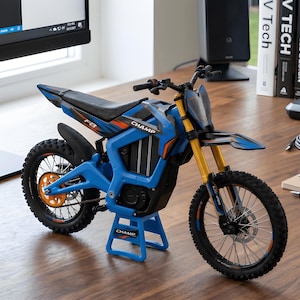 Puede incluir: Una moto de cross de juguete azul y negra con detalles en naranja y amarillo. La moto de juguete tiene la palabra "CHAMP" impresa en el lateral y en el soporte. Los neumáticos son negros con radios plateados.