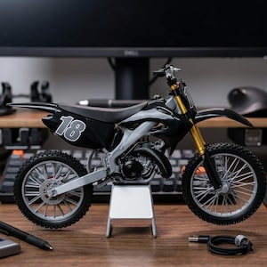 May include: A black and silver toy dirt bike with the number 18 on the side. The motorcycle has gold-colored front forks and is displayed on a small stand. The tires have a detailed tread pattern.