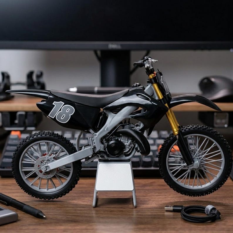 May include: A black and silver toy dirt bike with the number 18 on the side. The motorcycle has gold-colored front forks and is displayed on a small stand. The tires have a detailed tread pattern.