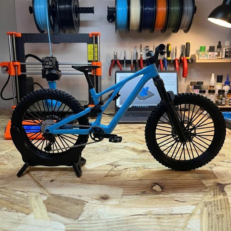 Puede incluir: Una maqueta de bicicleta de monta&ntilde;a azul con neum&aacute;ticos y componentes negros. La bicicleta se exhibe en un soporte negro, mostrando su dise&ntilde;o intrincado. El fondo incluye una impresora 3D y herramientas.