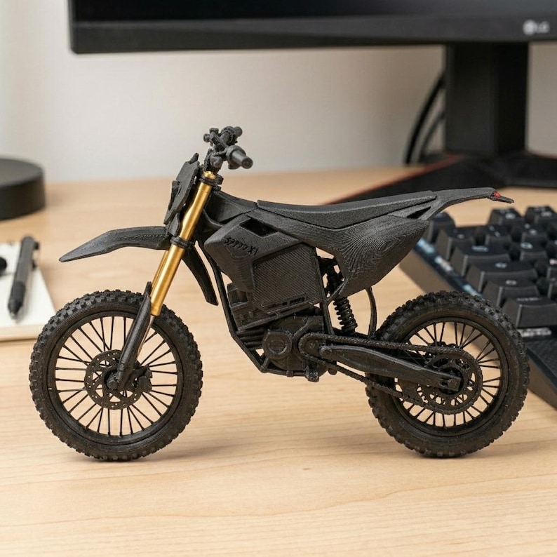 Puede incluir: Modelo en miniatura de moto de cross negra y dorada. El modelo presenta un dise&ntilde;o detallado con un asiento texturizado, neum&aacute;ticos con tacos y horquillas delanteras doradas. El texto "SPPPX XL" es visible.