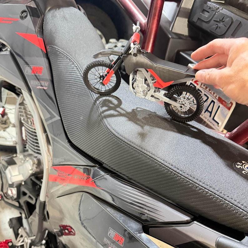 Puede incluir: Una maqueta de moto de cross negra y roja con una matr&iacute;cula que dice "AL" se coloca sobre un asiento de motocicleta. El modelo tiene detalles, incluyendo ruedas, un asiento y manillar. El asiento es negro con un patr&oacute;n texturizado.