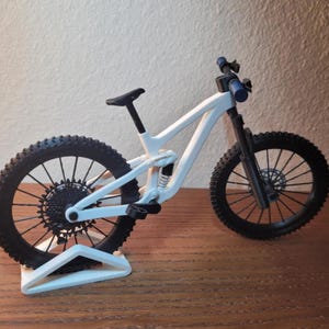 May include: A white mountain bike model with black tires and components. The bike has a suspension system and is displayed on a white stand. The handlebars are blue. The bike is on a wooden surface.