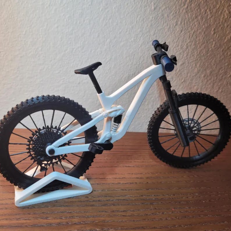 May include: A white mountain bike model with black tires and components. The bike has a suspension system and is displayed on a white stand. The handlebars are blue. The bike is on a wooden surface.