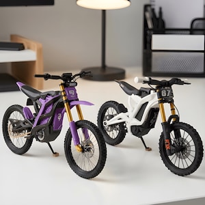 Op de afbeelding: Twee miniatuur elektrische crossmotoren, één paars en één wit, met goudkleurige voorvorken, zwarte spaakwielen en zwarte zadels. De witte motor heeft het nummer 67 op de voorplaat, terwijl de paarse motor een uniek logo op de voorzijde heeft.