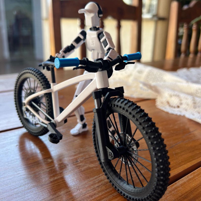 May include: A white and black mountain bike with blue handlebars. The bike has black tires and a white frame. A white and black action figure is posed behind the bike.
