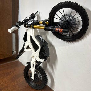 Puede incluir: Una minimoto blanca y negra con detalles dorados, montada en una pared. Tiene neum&aacute;ticos negros, un asiento negro y un manillar blanco y negro. La rueda delantera est&aacute; en el suelo, mientras que la trasera est&aacute; elevada.