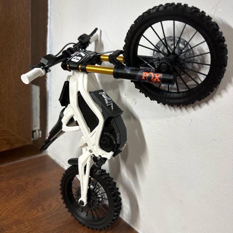 Puede incluir: Una minimoto blanca y negra con detalles dorados, montada en una pared. Tiene neum&aacute;ticos negros, un asiento negro y un manillar blanco y negro. La rueda delantera est&aacute; en el suelo, mientras que la trasera est&aacute; elevada.