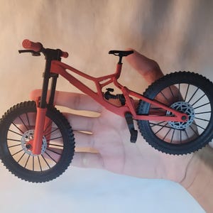 K&ouml;nnte beinhalten: Ein rotes und schwarzes Miniatur-Mountainbike mit schwarzen Reifen und einem schwarzen Sattel. Das Fahrrad hat ein Federungssystem und Scheibenbremsen. Der Lenker ist rot mit schwarzen Griffen.