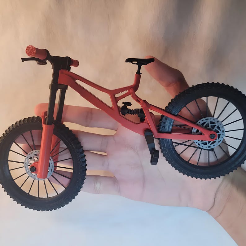 K&ouml;nnte beinhalten: Ein rotes und schwarzes Miniatur-Mountainbike mit schwarzen Reifen und einem schwarzen Sattel. Das Fahrrad hat ein Federungssystem und Scheibenbremsen. Der Lenker ist rot mit schwarzen Griffen.