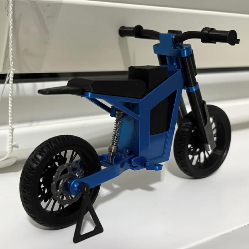 Puede incluir: Modelo de motocicleta azul y negro en miniatura. El modelo presenta neum&aacute;ticos negros, un asiento negro y suspensi&oacute;n de resorte plateada. La motocicleta se exhibe sobre una superficie blanca. R&eacute;plica detallada.