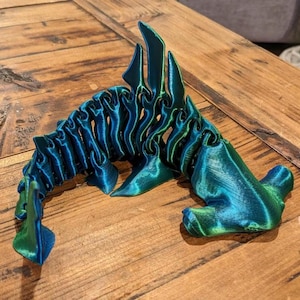 May include: A 3D-printed, articulated hammerhead shark figurine. The shark is a gradient of blue and green, with a textured, ridged spine and fins. The figurine is posed on a wooden surface.