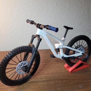 May include: A white and black mountain bike model with blue handlebar grips. The bike has black tires, a black seat, and a silver disc brake. It is displayed on a red stand, showcasing its design and features.