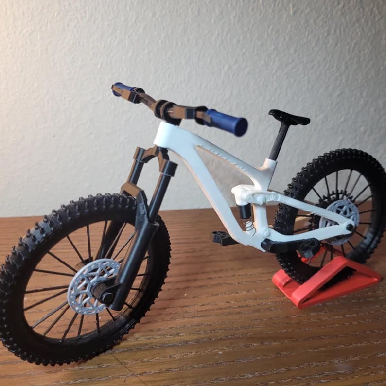 May include: A white and black mountain bike model with blue handlebar grips. The bike has black tires, a black seat, and a silver disc brake. It is displayed on a red stand, showcasing its design and features.