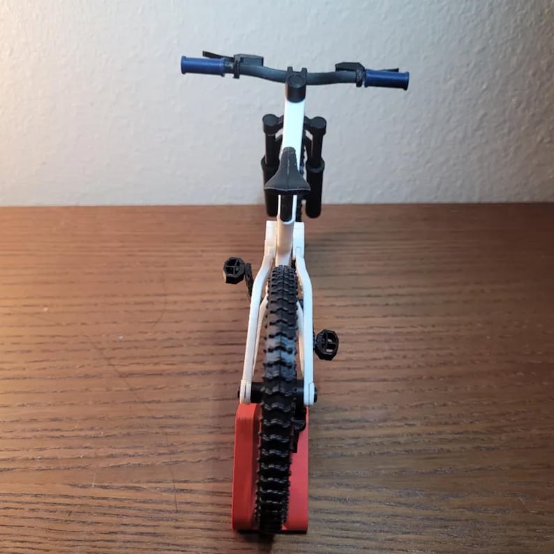 May include: A white and black mountain bike model with blue handlebars and black tires. The bike is mounted on a red stand, showcasing its design. The bike's frame is white, and the tires have a detailed tread pattern.