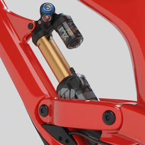 May include: Close-up of a red mountain bike frame with a gold and black Fox Factory shock absorber. The shock absorber has blue and red adjustment dials. The frame has black hardware and a glossy finish.