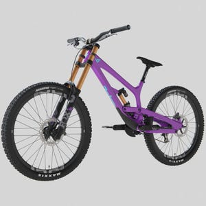 Könnte beinhalten: Ein lila Mountainbike mit schwarzen Reifen und einem schwarzen Sattel. Das Fahrrad hat goldfarbene Federgabeln und einen schwarzen Lenker. Auf den Rädern steht das Wort "MAXXIS" auf den Reifen. Der Rahmen hat ein Dreieckslogo.