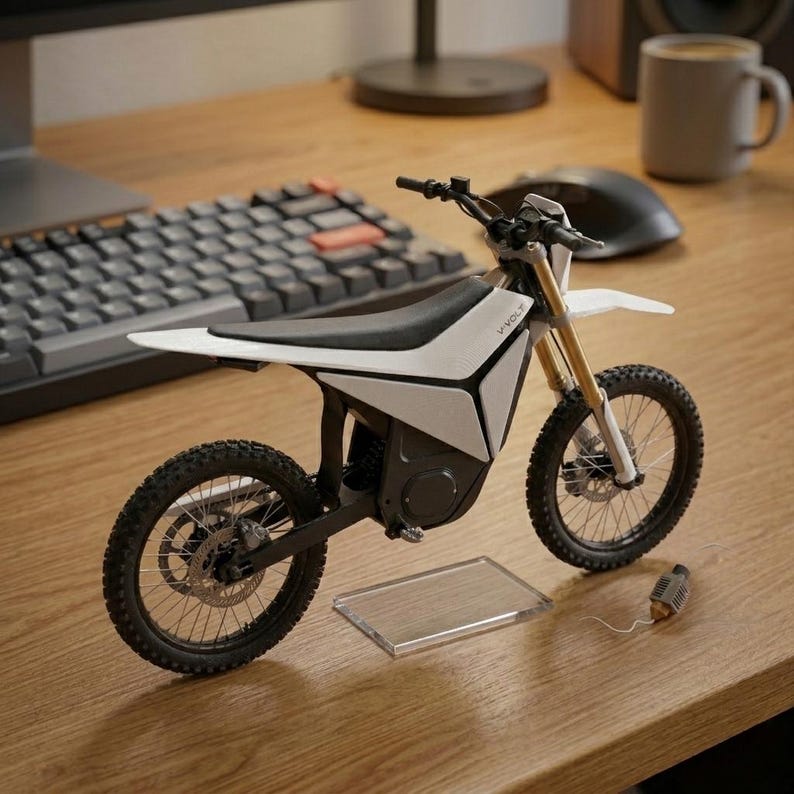 Op de afbeelding: Een wit en zwart elektrisch crossmotor model met de tekst "V-Volt" aan de zijkant. Het model heeft zwarte banden, gouden vering en een doorzichtige display standaard. De motor staat op een houten oppervlak.