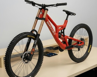 Puede incluir: Una bicicleta de montaña roja Santa Cruz V10 con cuadro de doble suspensión, horquillas de suspensión delanteras doradas, manillar negro y neumáticos negros con tacos y la marca Maxxis. Incluye amortiguador trasero y frenos de disco.