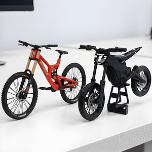May include: A red Santa Cruz mountain bike model with tan front forks and black tyres, positioned next to a black electric dirt bike model featuring a number one plate on the handlebars and resting on a small black stand.