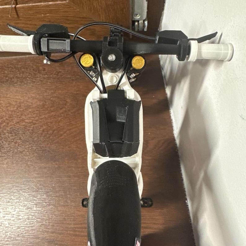 May include: A close-up view of a white and black scooter. The scooter has a black handlebar, a black front wheel, and yellow accents. The handlebars have white grips. The scooter appears to be made of plastic.