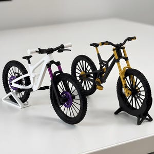 May include: Two miniature mountain bike models displayed on stands. One bike features a white frame with purple accents on the front fork and disc brakes. The second bike has a black frame with gold accents on the front fork and pedals.